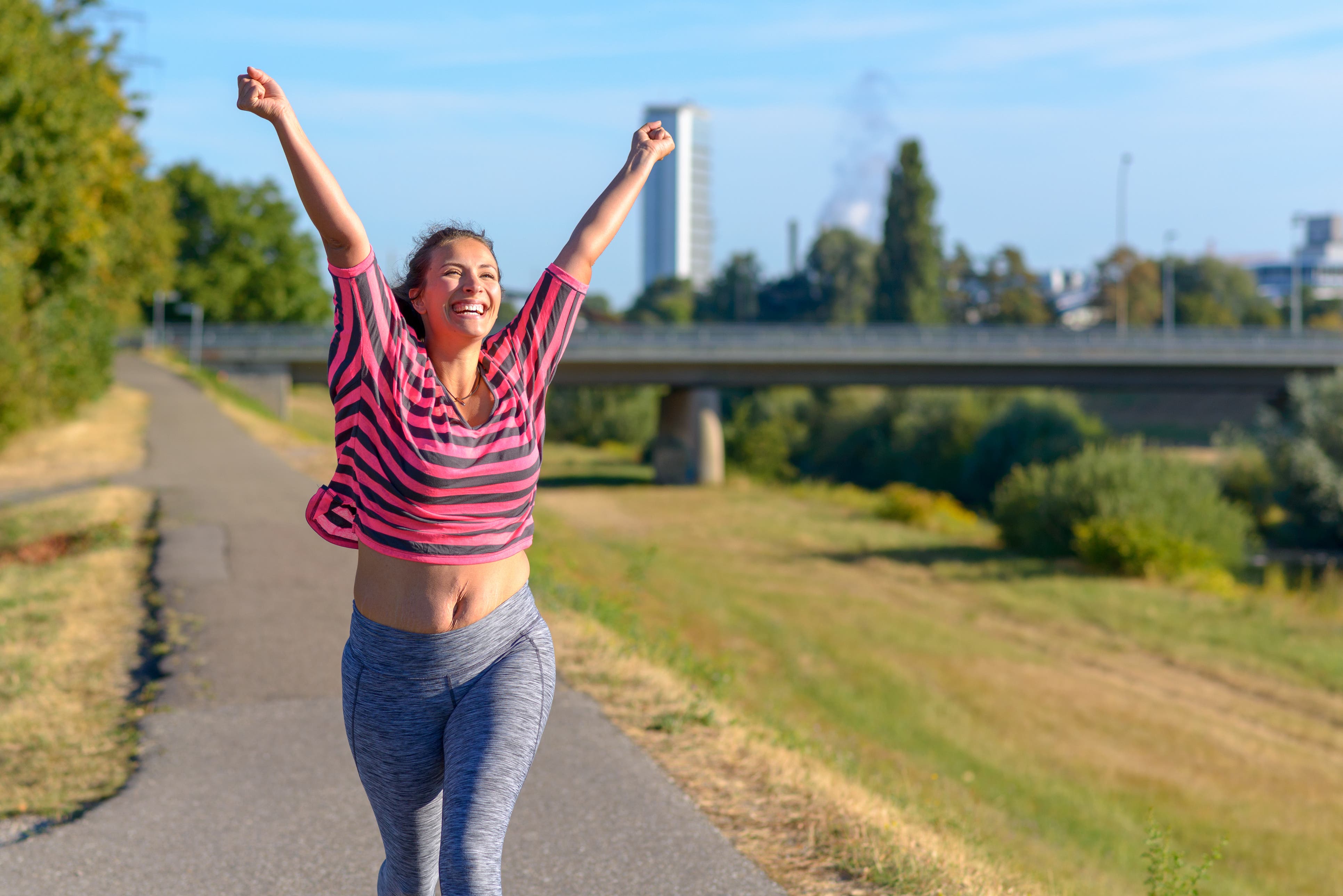 A young fit woman is smiling and raising her arms in the air as she walks outdoors