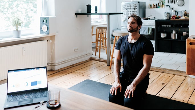 A man is on his knees in a living room on a black yoga mat, he is meditating for energy.