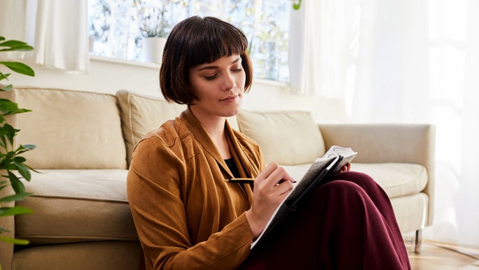 Contemplative woman sitting on the lounge room floor with her back up against the couch writing in her journal