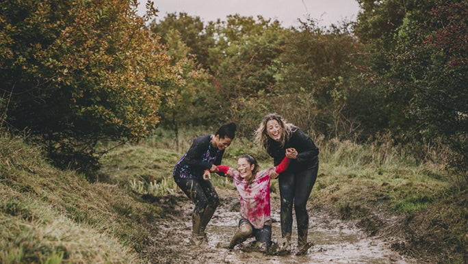 Three women are laughing in the mud, two of them are helping their friend up. It looks like they are doing an obstacle course. The perfect resilience skills training activity.