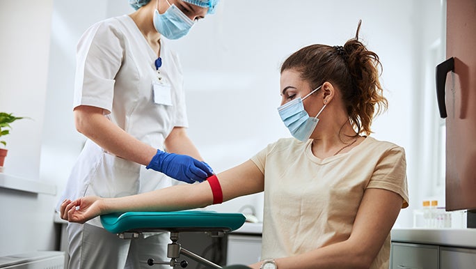 A masked woman is having a blood test. She has a red band around her arm.