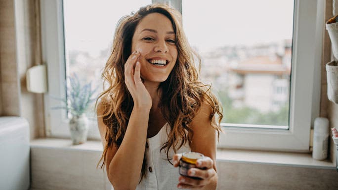 Smiling young woman applying face cream in the morning