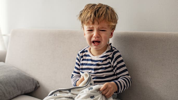 A little boy is crying on the couch, he is holding a blanket.