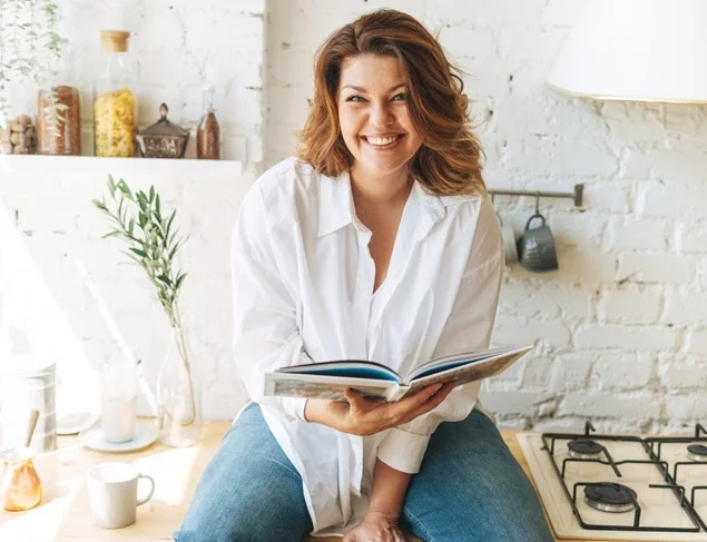 Happy young woman in blue jeans and white shirt reading cooking book in the home kitchen