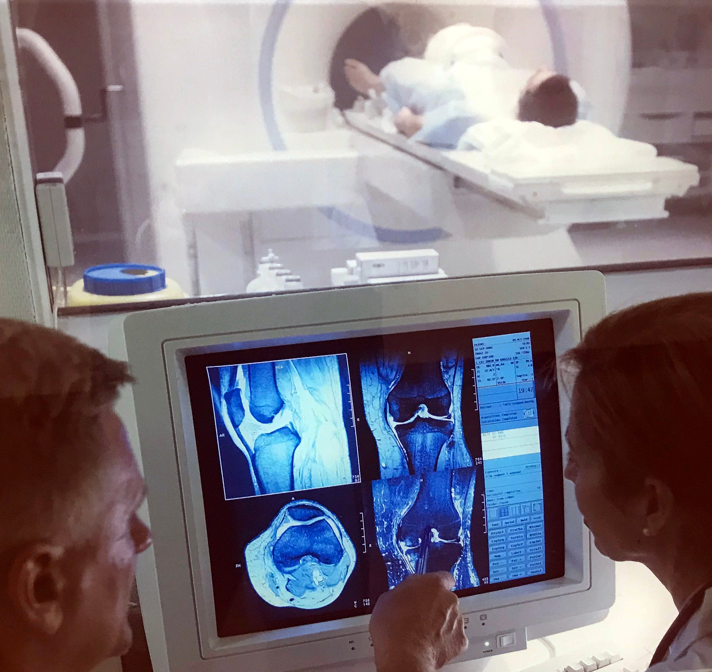 In the foreground, two medical professionals look at MRI images on a computer monitor, while in the background a patient lies on the MRI scanner bed