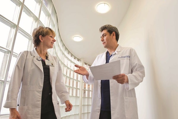 Two medical professionals in white lab coats, a male and a female - the male is holding a piece of paper and gesturing as they chat in a brightly-lit corridor