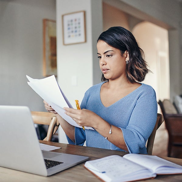 A young woman with dark hair is sitting at a table in front of a laptop and an open book, holding papers and a pen 