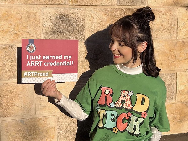 Smiling R.T. wearing a green Rad Tech T-shirt, holding a sign that says, 