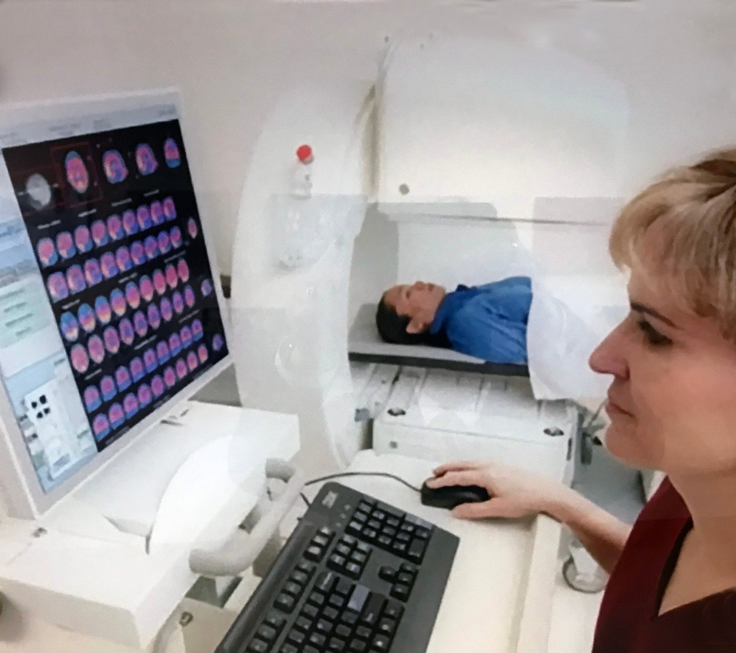 In the foreground, a nuclear medicine technologist looks at images on a computer monitor, while a patient lies on the scanner bed in the background