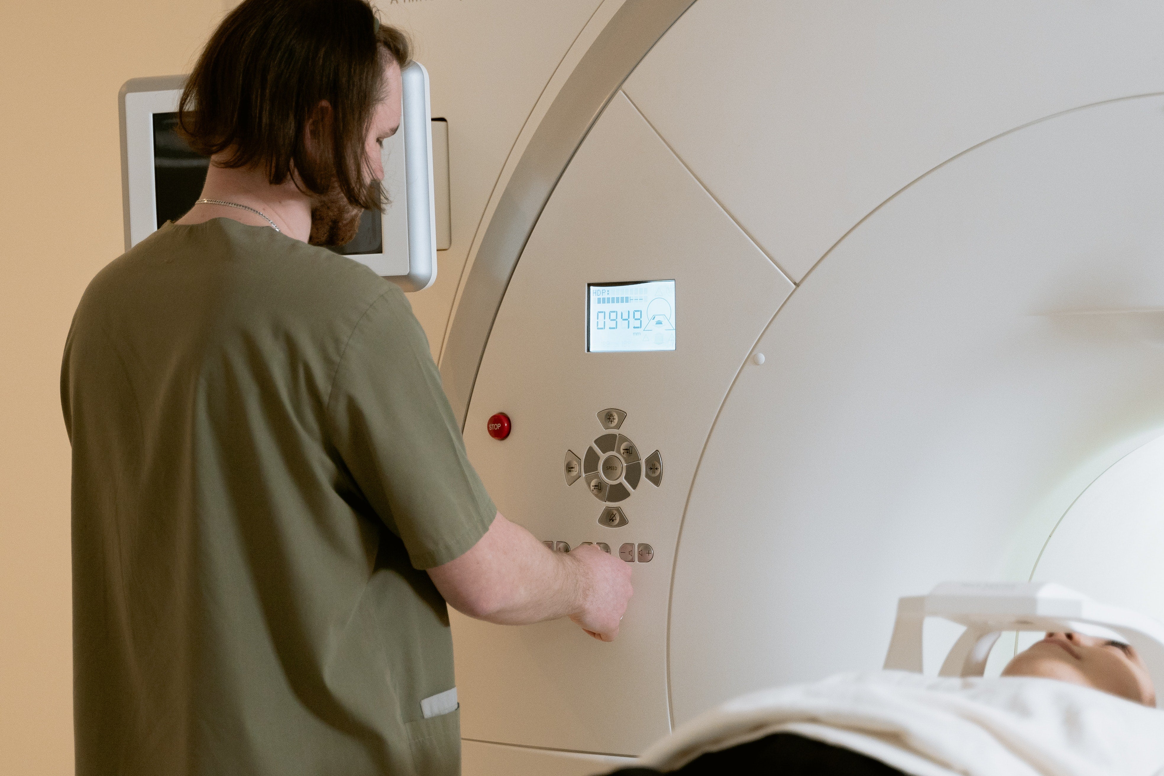 A male R.T. in scrubs with his back to the camera starting a CT scanner with a patient on the scanner bed