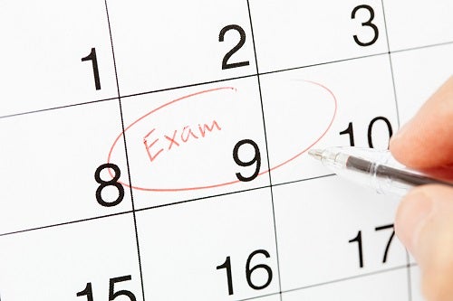 A hand holding a pen above a paper calendar with "Exam" written and circled on the ninth of the month 