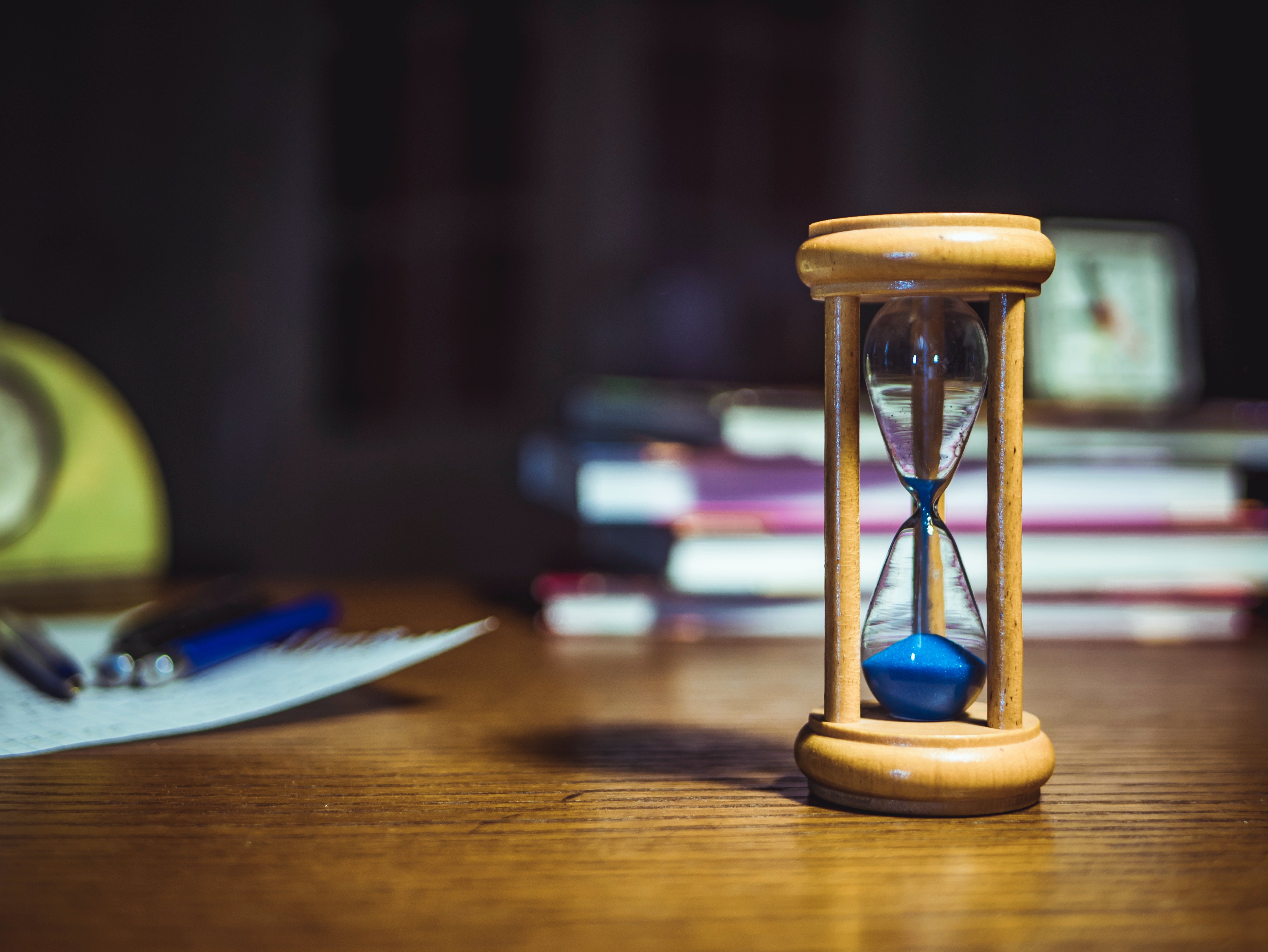 A wooden hourglass with blue sand in the foreground, with a blurry stack of books and a clock in the background