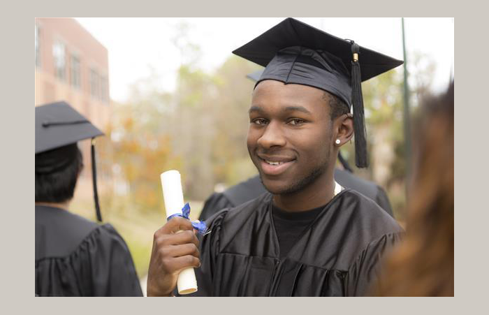 A smiling young man in a graduation cap and gown, holding a rolled up certificate that's tied with a blue ribbon