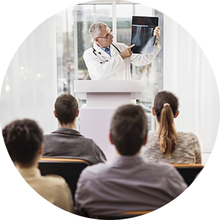 Round photo of a male radiography instructor wearing a lab coat holding up an X-ray in the background; the backs of four students heads in the foreground