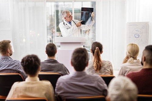 Speaker in a room full of students, wearing a lab coat and holding and pointing to an X-ray