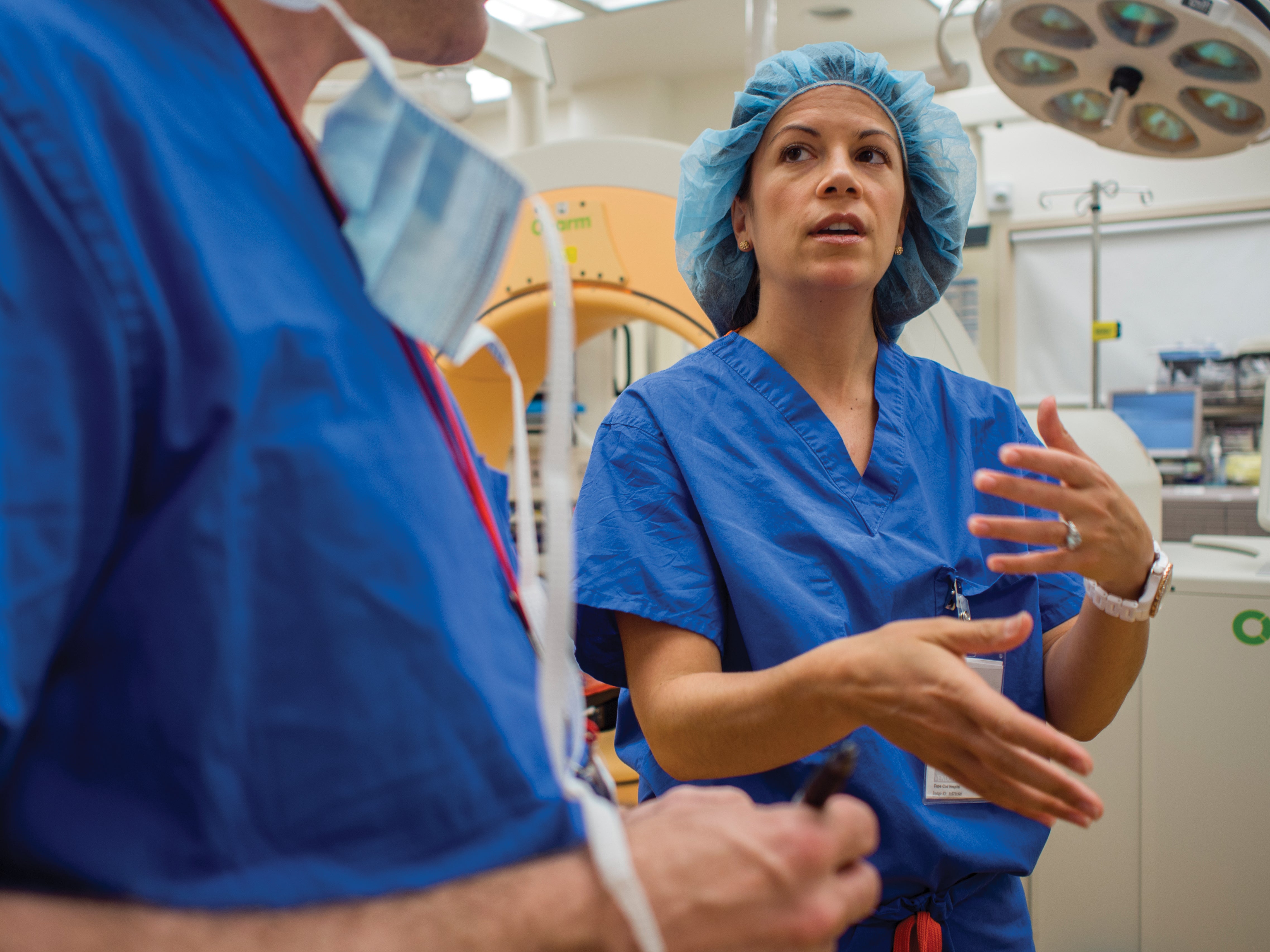 Two R.T.s in blue scrubs talking and gesturing in front of a CT scanner