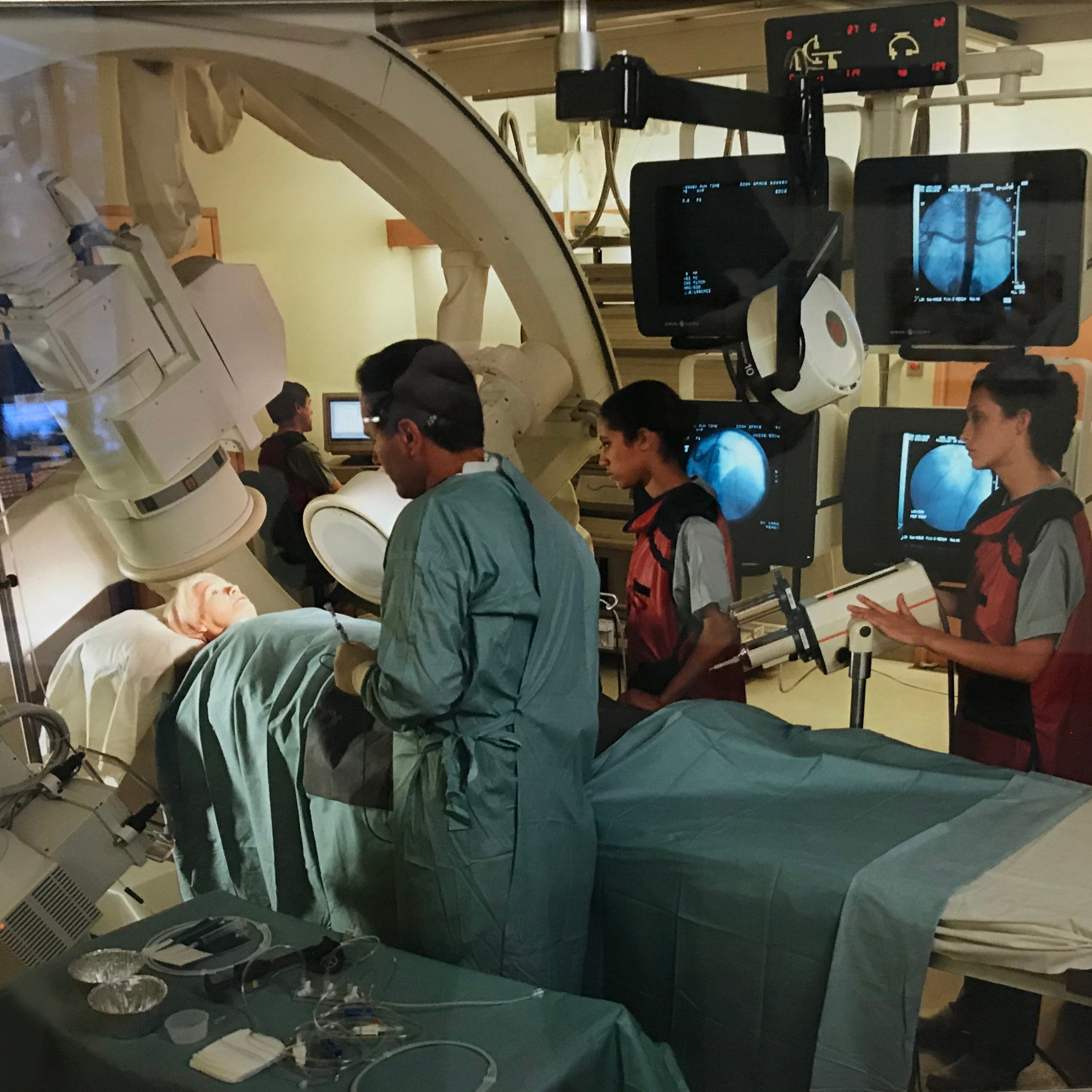 A patient is laying on a scanner bed in an imaging suite, while four medical professionals in surgical scrubs and radiation shielding work around her