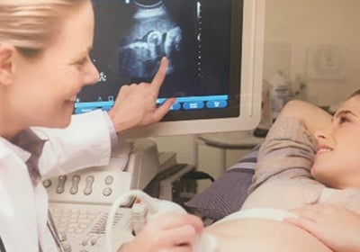 A pregnant woman is laying on an exam table while a smiling sonographer holds a transducer to the patient's belly with her right hand, and points at the image on the monitor with her left hand.