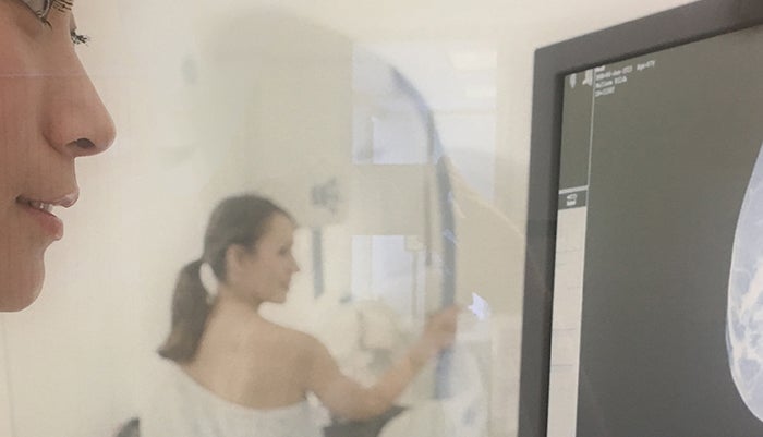 A mammographer looks at an image on a monitor in the foreground while a patient stands at a mammography machine in the background with her back to the camera.