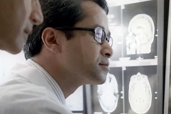 Medical professional with dark hair and glasses is looking at medical imaging scans while another medical professional looks over his shoulder.