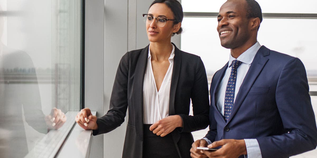 two diverse business professionals, both smiling, looking out of an office building window together?auto=format&q=75