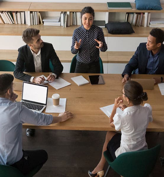 A woman standing and speaking to four team members around a conference table.?auto=format&q=75