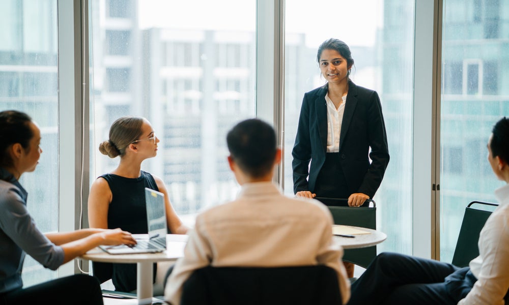 Indian Asian woman stands up in front of her diverse team and is leading a meeting, training or presentation in their office during the daytime. .jpeg