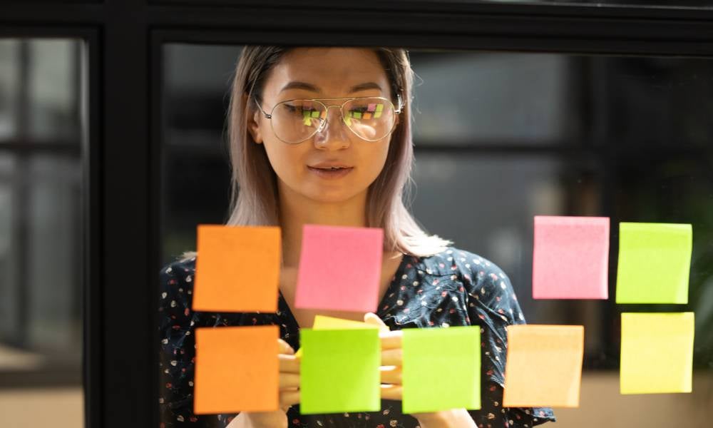 Serious focused young female coach teacher student asian business woman working on project strategy plan writing target tasks creative ideas on sticky post it notes on glass scrum board office wall.jpeg