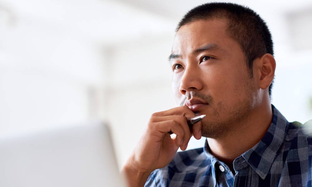 Man deep in thought with a hand on his chin while working on a laptop alone at a desk in a modern office (1).jpg