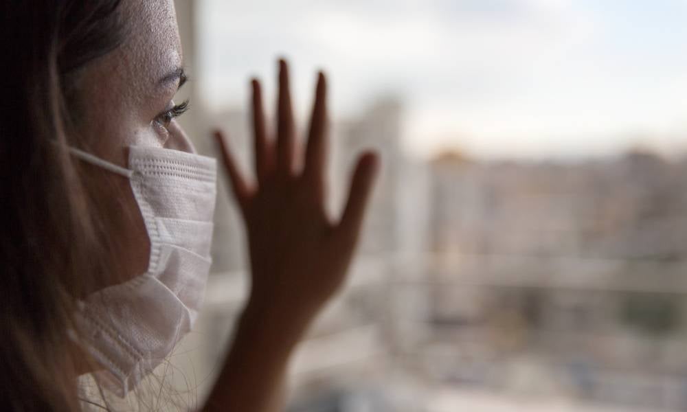 Woman wearing a face mask looks outside her window hand on glass.jpeg
