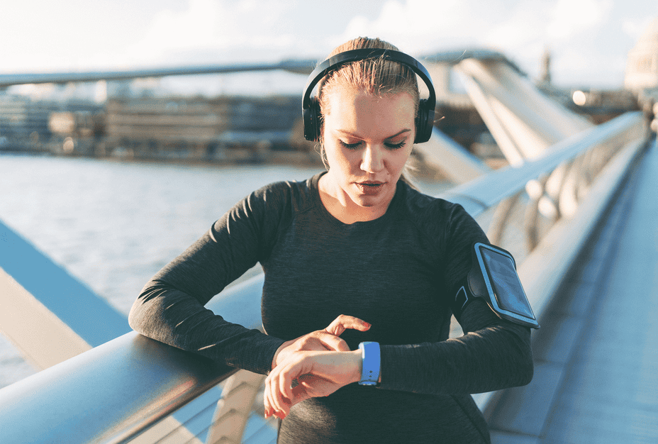 Woman pauses mid-run to read her smart watch, while wearing headphones and smart phone on her arm