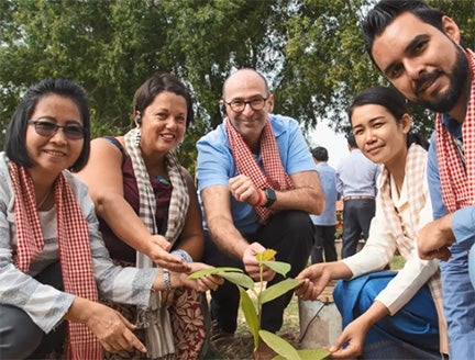 A group of 5 both men and women bent down smiling at the camera whilst touching a plant
