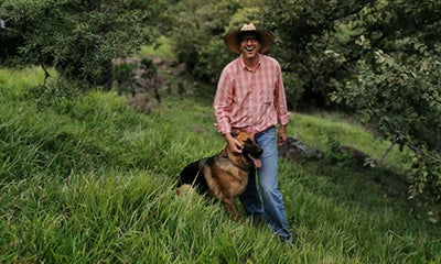 Hombre caminando por un campo sonriendo a la cámara mientras toca a su perro