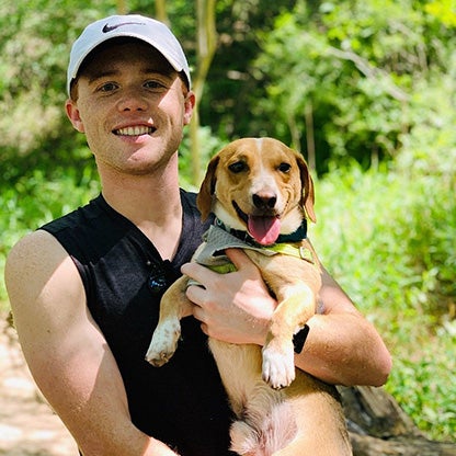 a young man smiling whilst holding a small dog in a forest