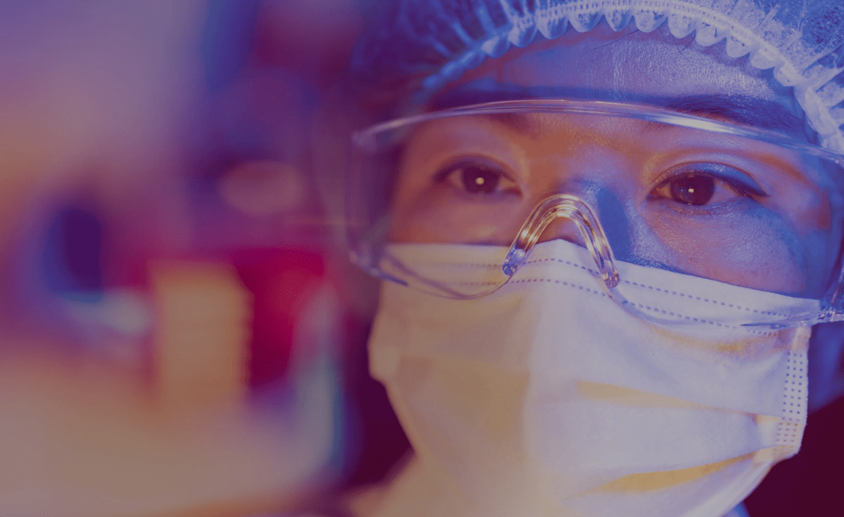 A scientist wearing a face mask, goggles, and a hair net looks at something out of focus in front of her