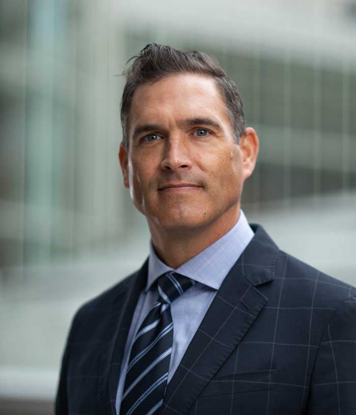 Nathan LeBrassuer, a man with brown hair, wears a dark blue windowpane suit, a light blue shirt, and a dark blue striped tie.