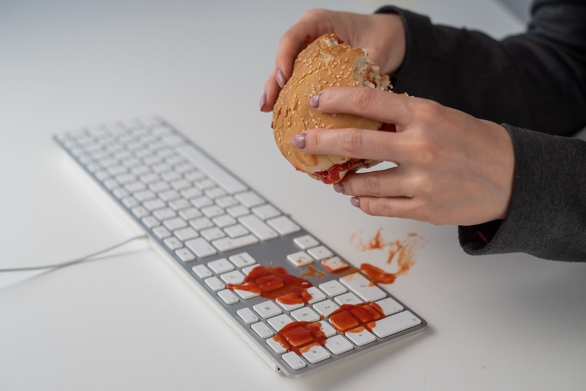 Hands hold a hamburger dripping ketchup over a keyboard covered with splatters of ketchup