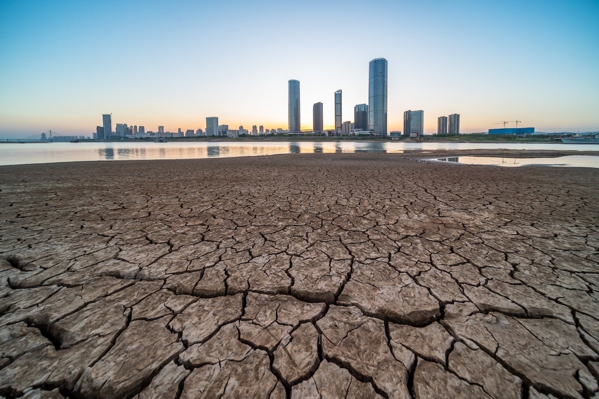 A dry, cracked ground in front of a body of water and city skyline at sunset