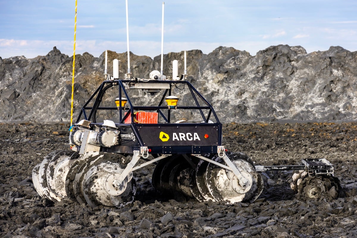 A rover that says ARCA in white lettering drives through a field of mine waste that looks like mud