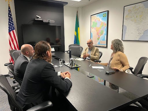 Two XPRIZE staff members sit across a black conference table from two USAID staff members. On the wall hangs maps of Brazil. There are also flags representing Brazil and the United States in the corners of the room.