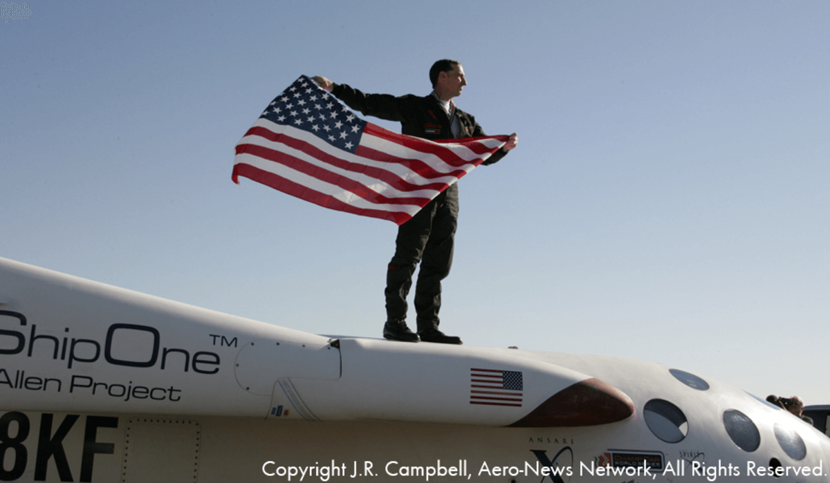 A man holding an American flag stands atop SpaceShipOne after the completion of the Ansari XPRIZE.