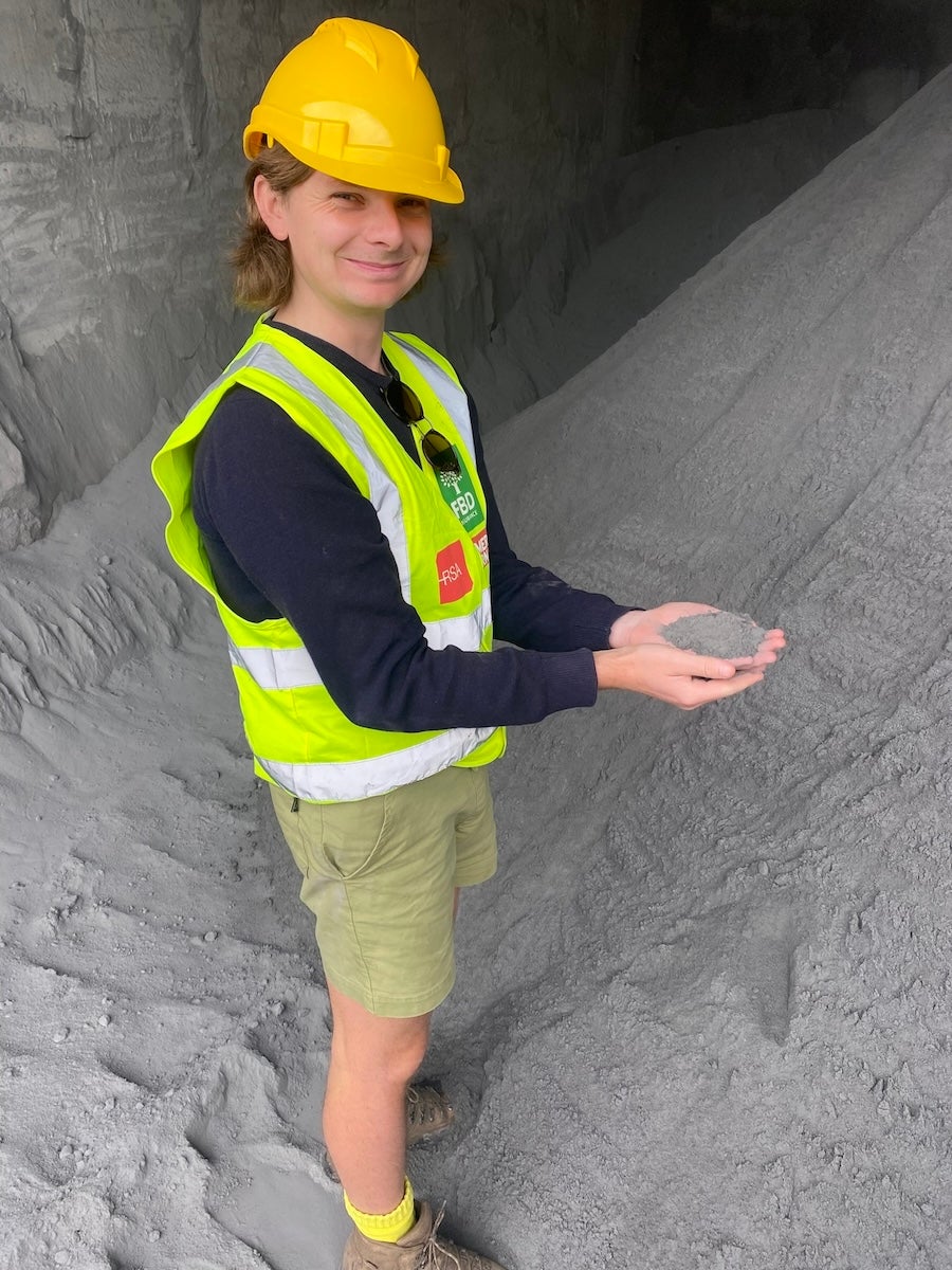 A man with shoulder length hair holds stands on a pile of ground-up concrete and holds some in his hands. He is wearing an orange-yellow hard hat and a fluorescent yellow safety vest.