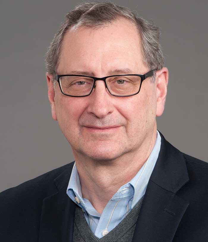 Stephen Kritchevsky, a man with brown hair, wears a blazer, gray sweater, and blue striped shirt on a gray background.