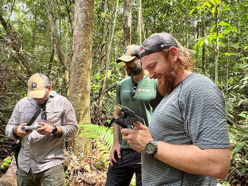 Three men in the rainforest look at screens on drone controllers.
