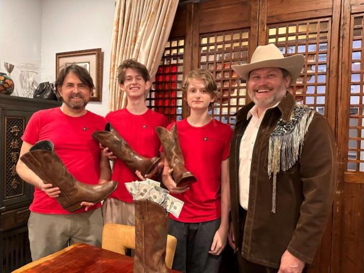 Chuck Brady, in a white cowboy hat and brown, fringed jacket, stands with the three members of AvaDynamics, wearing matching red shirts. AvaDynamics members are each holding a custom XPRIZE-branded cowboy boot. In front of them, another boot from the set is on a table with dollar bills sticking out of the top.