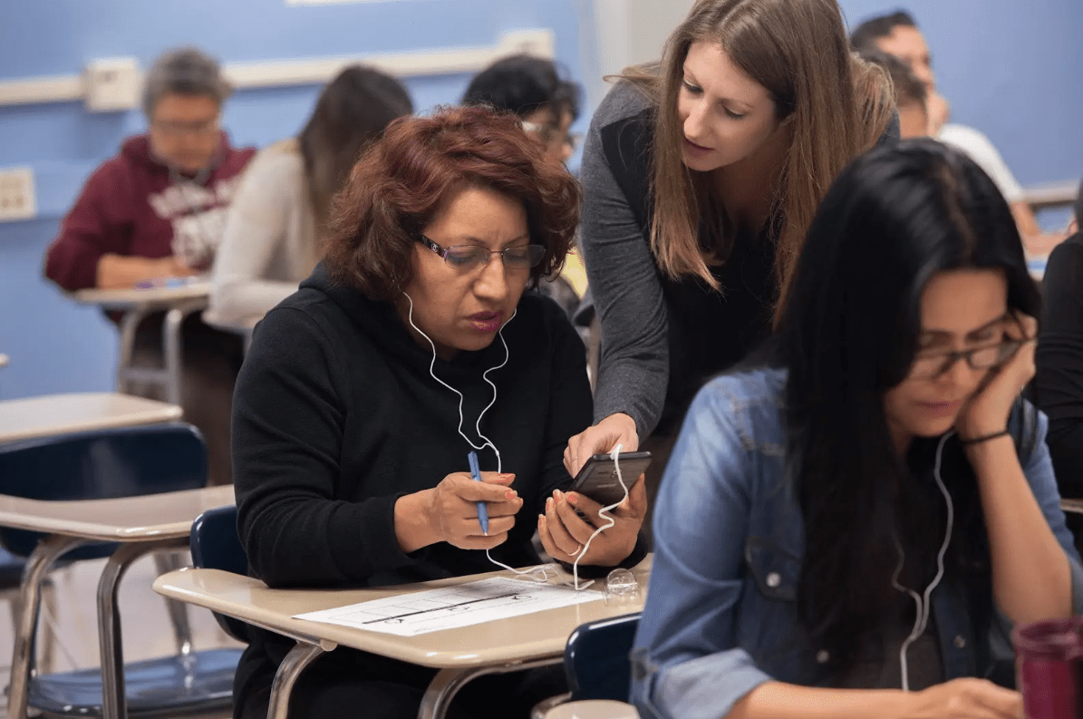 In a classroom of adults listening with headphones, a teacher leans in and helps a student