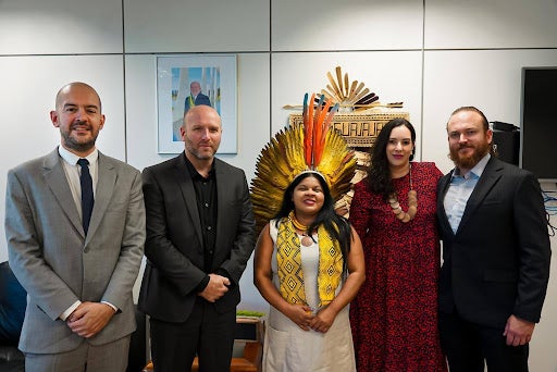 A line of five people in formal attire. In the middle is Minister Sonia Guajajara, who is wearing a traditional indigenous headdress and other clothing items