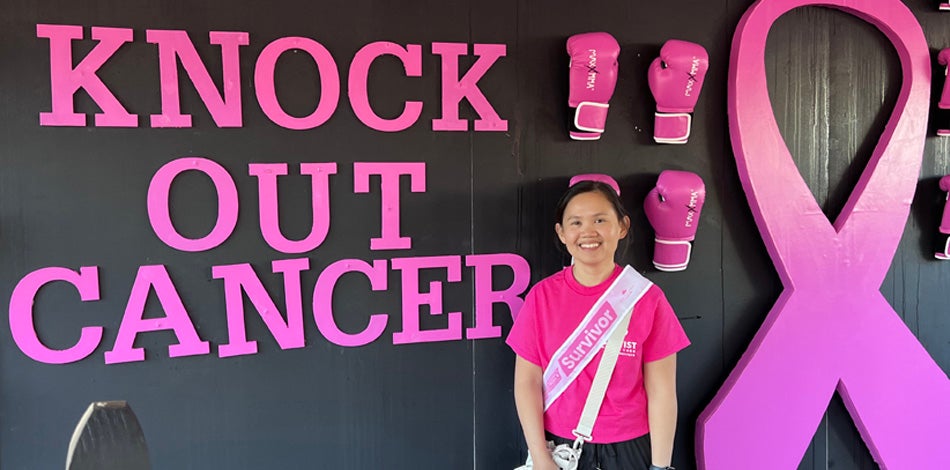 Young female, Asian wears pink and stands in front of words that say Knock Out Cancer