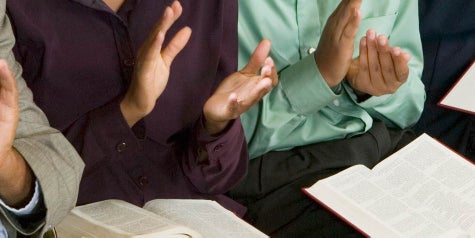 Church members sitting in a pew clapping hands to worship with Bibles in lap.