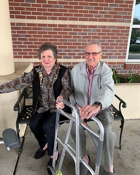 Marrianne and Earl, elder white, married couple, sit on bench together smiling 